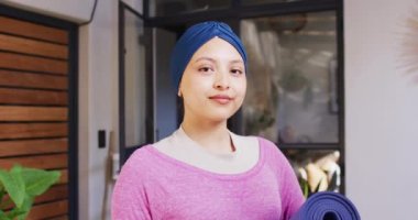 Evdeki oturma odasında kameraya gülümseyen mavi tesettürlü mutlu çift ırklı kadının video portresi. Mutluluk, sağlık, kapsayıcılık ve ev hayatı.
