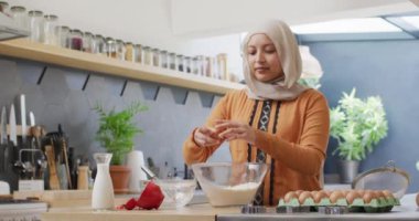 Mutfakta tesettürlü mutlu melez kadın videosu. Karıştırma kabına yumurta kırıyor. Mutluluk, sağlık, kapsayıcılık ve ev hayatı.