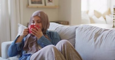 Türbanlı düşünceli bir kadının evdeki oturma odasındaki kanepede kahve içerken çekilmiş videosu. Mutluluk, rahatlama, kapsayıcılık ve ev hayatı..