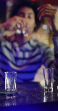 Vertical video of drunk african american man sitting at a bar drinking shots. Drinking, going out, inclusivity and alcohol concept.