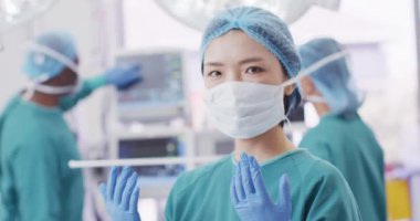 Video portrait of asian female surgeon in cap, gloves and face mask in operating theatre, copy space. Hospital, medical and healthcare services.