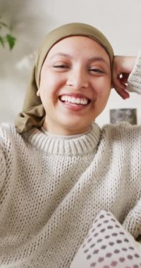 Vertical video of smiling biracial woman in hijab making video call talking to camera. Happiness, communication, relaxation, inclusivity and domestic life.