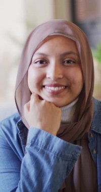 Vertical video portrait of happy biracial woman in brown hijab smiling to camera. Happiness, relaxation, inclusivity and domestic life.