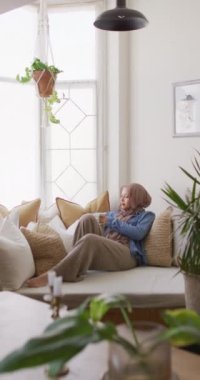 Vertical video of thoughtful biracial woman in hijab relaxing on sofa drinking coffee in living room. Happiness, relaxation, inclusivity and domestic life.