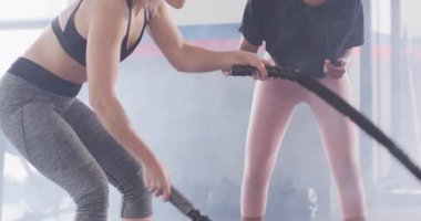 Video of diverse female fitness trainer with stopwatch and woman battling ropes working out at a gym. Exercise, fitness and healthy lifestyle.