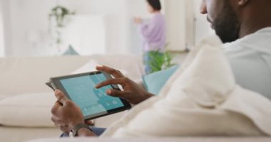 Diverse couple sitting on couch, using tablet and walking in living room. Spending quality time at home concept.