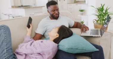 Diverse couple lying on couch and using laptop in living room. Spending quality time at home concept.