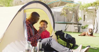 Happy african american father and his son sitting in tent and using tablet in garden. Spending quality time at home.