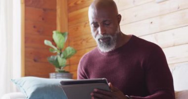 Happy senior african american man spending time in log cabin, sitting on sofa and using tablet. Free time, domestic life and nature concept.