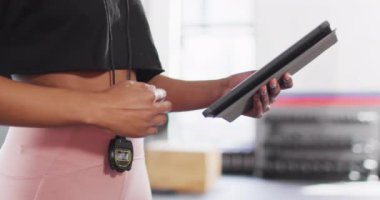 Video of midsection of african american female fitness trainer using stopwatch and tablet at a gym. Exercise, fitness and healthy lifestyle.