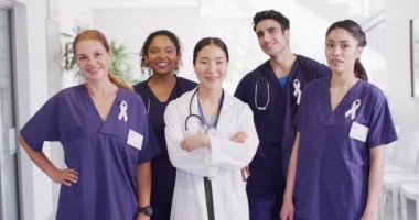 Video portrait of diverse group of happy doctors wearing cancer ribbons smiling in hospital corridor. Hospital, medical and healthcare services.