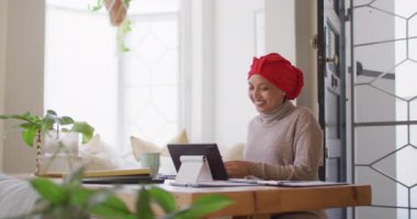 Video of happy biracial woman in hijab sitting at desk making tablet video call at home, copy space. Working from home, communication, inclusivity and lifestyle.