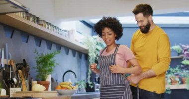 Video of happy diverse couple putting on apron and talking in kitchen at home, with copy space. Happiness, inclusivity, free time, togetherness and domestic life.