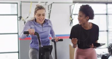 Video of diverse female fitness trainer and woman on exercise bike bumping fists at a gym. Exercise, fitness and healthy lifestyle.