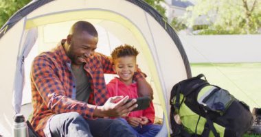Happy african american father and his son sitting in tent and using smartphone in garden. Spending quality time at home.