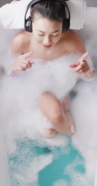 Vertical video of smiling biracial woman with headphones in bathtub in bathroom. Health and beauty, leisure time, domestic life and lifestyle concept.