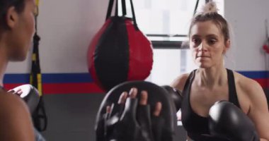 Video of diverse female coach in pads and smiling woman in boxing gloves boxing training at a gym. Exercise, fitness and healthy lifestyle.