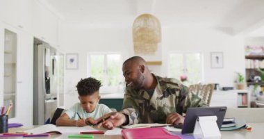 Happy african american male soldier and his son sitting at table, working and doing homework. Spending quality time together, army and patriotism concept.
