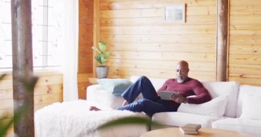 Happy senior african american man spending time in log cabin, sitting on sofa and using tablet. Free time, domestic life and nature concept.