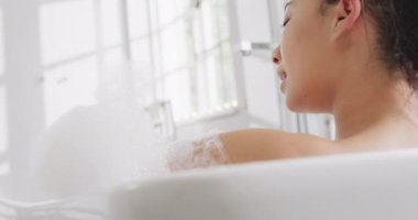 Video of smiling biracial woman lying in bathtub in bubble bath in bathroom. Health and beauty, leisure time, domestic life and lifestyle concept.