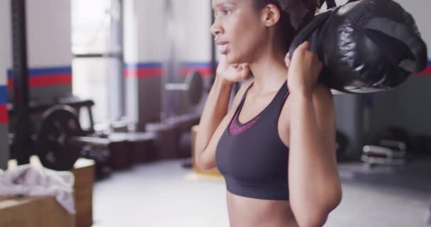 決定されたアフリカ系アメリカ人女性がジムで砂袋の重さを持つスクワットをしているビデオ フィットネス 健康的なライフスタイル — ストック動画