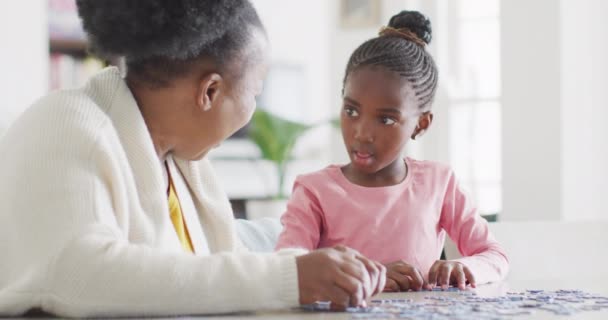 幸せなアフリカ系アメリカ人の孫娘と祖母が家でジグソーパズルをして スペースをコピーします 家族と国内生活 — ストック動画