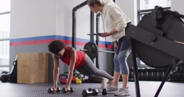 Video of diverse female fitness trainer instructing woman raising dumbbells working out at a gym. Exercise, fitness and healthy lifestyle.