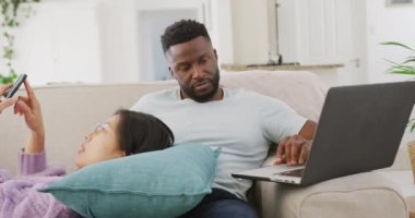 Diverse couple lying on couch and using laptop in living room. Spending quality time at home concept.