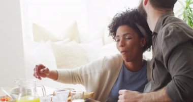 Video of happy diverse couple eating breakfast and talking at table. Happiness, inclusivity, free time, togetherness and domestic life.
