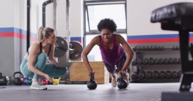 Video of diverse female fitness trainer instructing woman doing push ups on kettlebells at gym. Exercise, fitness and healthy lifestyle.