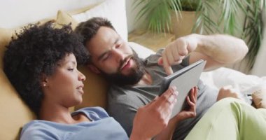 Video of happy diverse couple relaxing at home, lying on bed using tablet. Happiness, communication, inclusivity, free time, togetherness and domestic life.