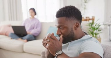 Diverse couple drinking coffee and using laptop in living room. Spending quality time at home concept.