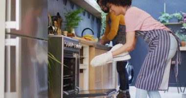 Video of happy diverse couple in aprons baking together in kitchen, using oven, with copy space. Happiness, communication, inclusivity, free time, togetherness and domestic life.