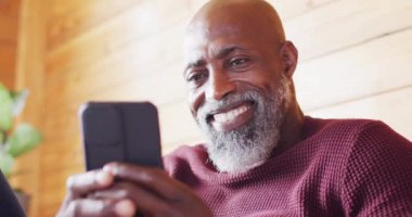 Happy senior african american man spending time in log cabin, sitting on sofa and using smartphone. Free time, domestic life and nature concept.