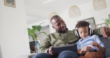 Happy african american man and his son sitting on sofa and using tablet. Spending quality time at home, childhood and family concept.