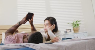 Happy diverse couple using smartphone and lying in bedroom. Spending quality time at home concept.
