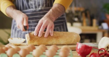 Video of midsection of caucasian man in apron slicing bread in kitchen, with copy space. Happiness, free time and domestic life.
