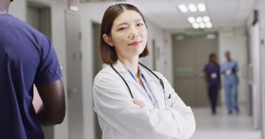 Video portrait of smiling asian female doctor standing in busy hospital corridor, copy space. Hospital, medical and healthcare services.