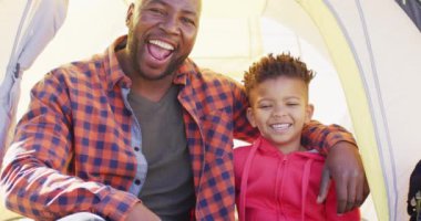 Portrait of happy african american father and his son sitting in tent and smiling in garden. Spending quality time at home.