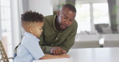 Happy african american man and his son sitting at table and reading braille. Spending quality time at home, childhood, blindness and family concept.