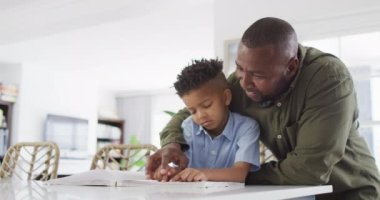 Happy african american man and his son sitting at table and reading braille. Spending quality time at home, childhood, blindness and family concept.