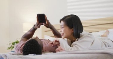 Happy diverse couple using smartphone and lying in bedroom. Spending quality time at home concept.