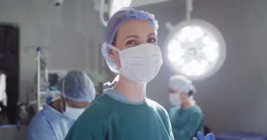 Video portrait of caucasian female surgeon in face mask smiling in operating theatre, copy space. Hospital, medical and healthcare services.