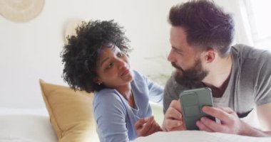 Video of happy diverse couple relaxing at home, talking and using smartphone. Happiness, communication, inclusivity, free time, togetherness and domestic life.
