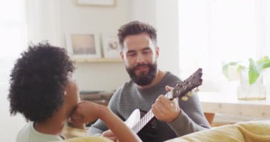 Video of rear view of happy diverse couple at home, man playing guitar, singing to woman, copy space. Happiness, inclusivity, free time, romance, music, togetherness and domestic life.