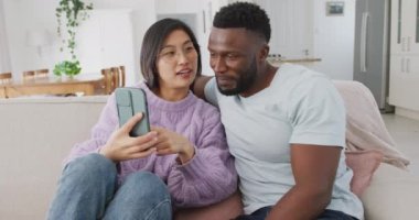 Diverse couple sitting on couch and using smartphone in living room. Spending quality time at home concept.