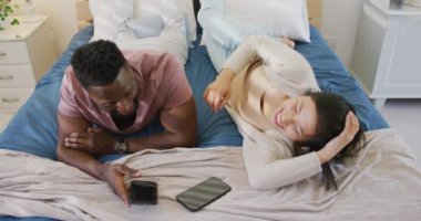 Happy diverse couple using smartphone and lying in bedroom. Spending quality time at home concept.