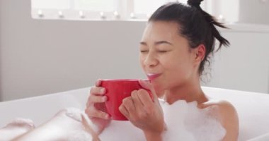 Video of portrait of smiling biracial woman sitting in bathtub in bubble bath with red cup of tea. Health and beauty, leisure time, domestic life and lifestyle concept.