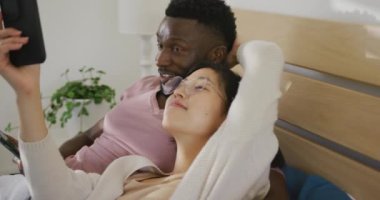 Happy diverse couple using smartphone and lying in bedroom. Spending quality time at home concept.