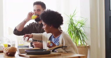 Video of happy diverse couple eating and drinking at breakfast table. Happiness, inclusivity, free time, togetherness and domestic life.